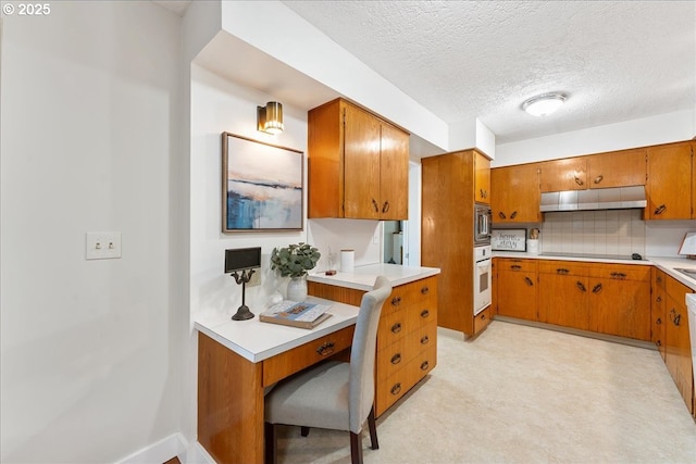 kitchen with under cabinet range hood, stainless steel microwave, brown cabinetry, oven, and a textured ceiling