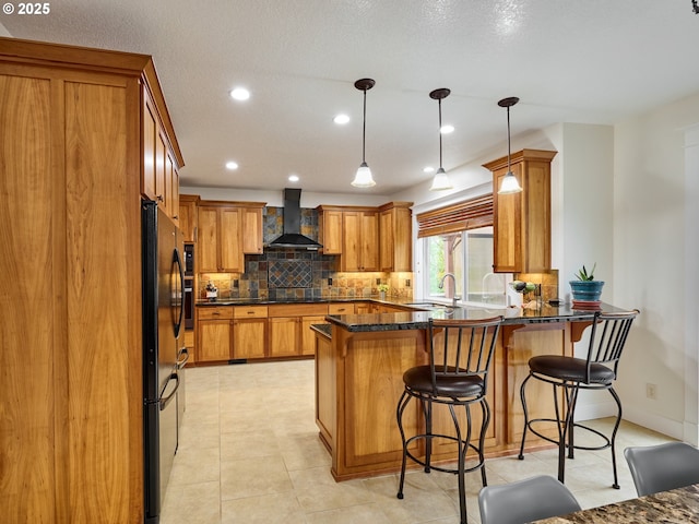 kitchen with a breakfast bar area, brown cabinetry, a peninsula, backsplash, and wall chimney range hood