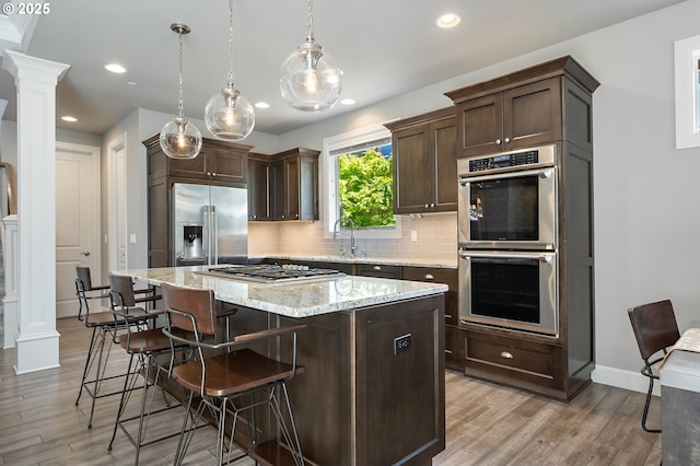 kitchen featuring decorative backsplash, decorative columns, dark brown cabinetry, a kitchen island, and appliances with stainless steel finishes