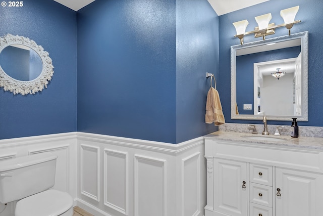 half bathroom with vanity, a decorative wall, toilet, and a wainscoted wall