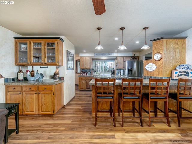 kitchen featuring light wood finished floors, glass insert cabinets, stainless steel fridge with ice dispenser, a breakfast bar area, and brown cabinetry