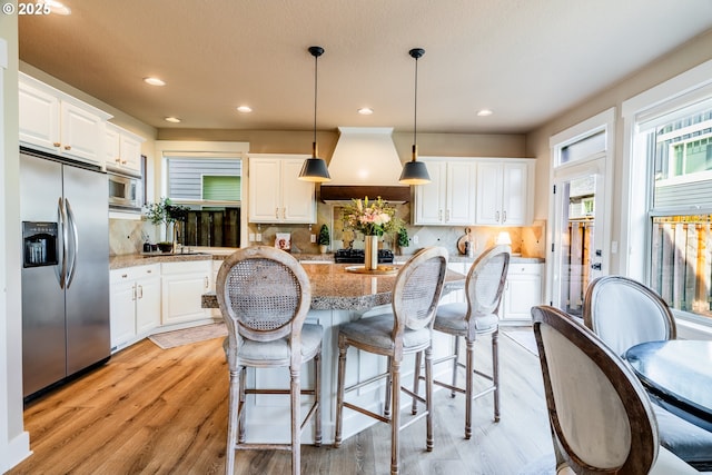 kitchen with custom exhaust hood, light stone counters, hanging light fixtures, stainless steel appliances, and white cabinets