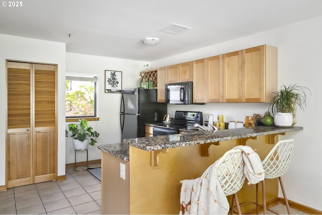 kitchen featuring black appliances, a peninsula, a kitchen bar, light tile patterned flooring, and dark stone countertops
