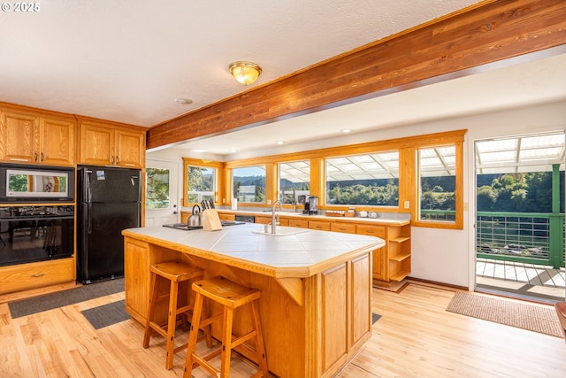 kitchen featuring a center island with sink, light wood-type flooring, tile counters, beamed ceiling, and black appliances