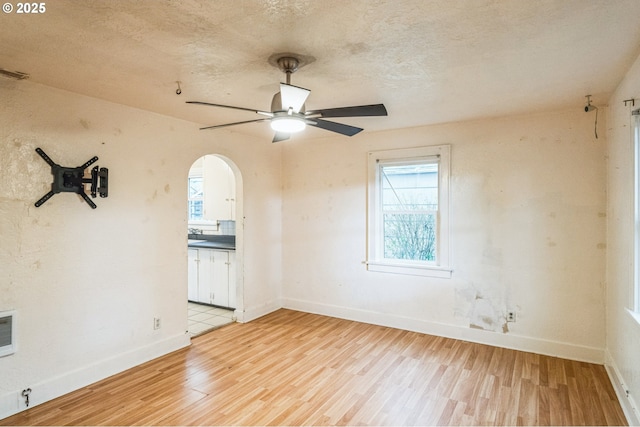 empty room featuring light wood-type flooring, a textured ceiling, arched walkways, baseboards, and ceiling fan