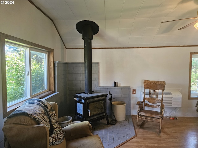 sitting room with a ceiling fan, a wood stove, vaulted ceiling, wood finished floors, and a wall mounted air conditioner