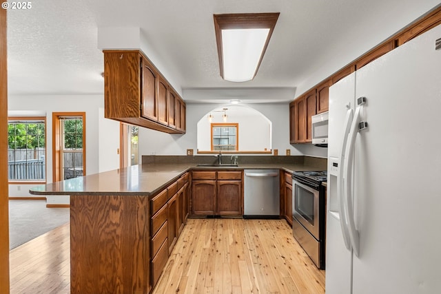 kitchen featuring stainless steel appliances, dark countertops, a peninsula, a sink, and light wood-style flooring