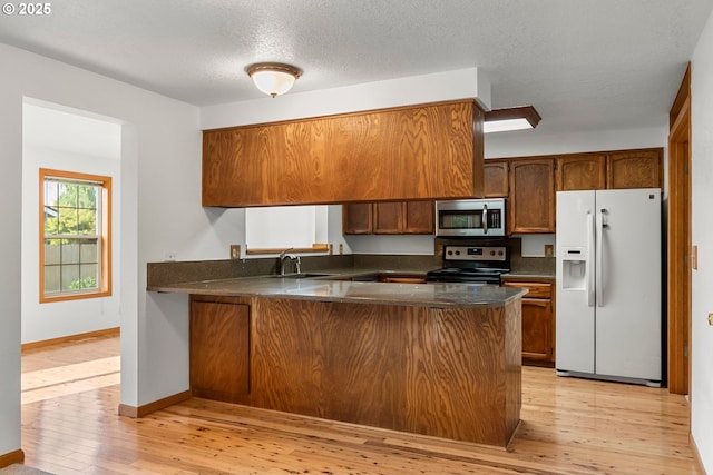 kitchen featuring appliances with stainless steel finishes, dark countertops, a peninsula, light wood finished floors, and brown cabinetry