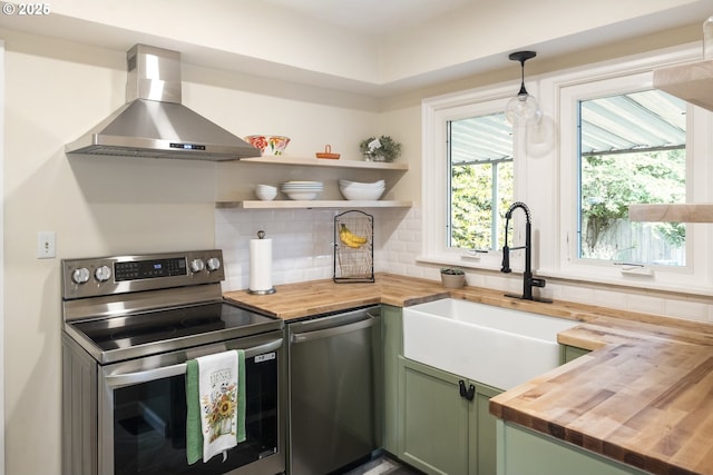 2535 N Baldwin St, Portland, OR 97217 - kitchen with butcher block countertops, stainless steel appliances, wall chimney exhaust hood, green cabinetry, and decorative backsplash