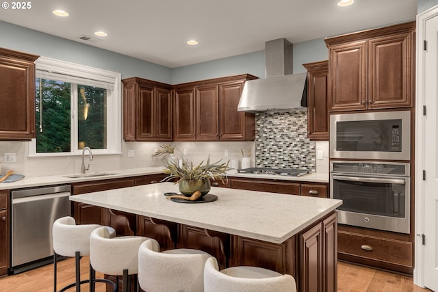 kitchen with appliances with stainless steel finishes, a kitchen bar, a center island, light wood-type flooring, and wall chimney exhaust hood