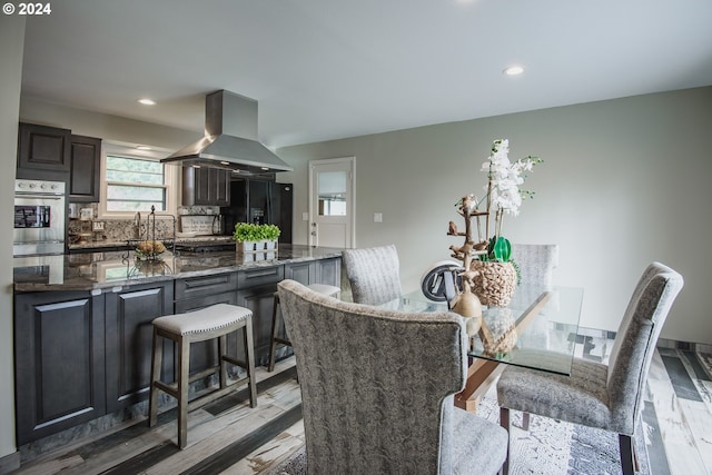 kitchen with a breakfast bar area, black refrigerator with ice dispenser, decorative backsplash, island range hood, and oven