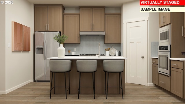 kitchen featuring wall oven, light wood-style floors, an island with sink, under cabinet range hood, and refrigerator with ice dispenser