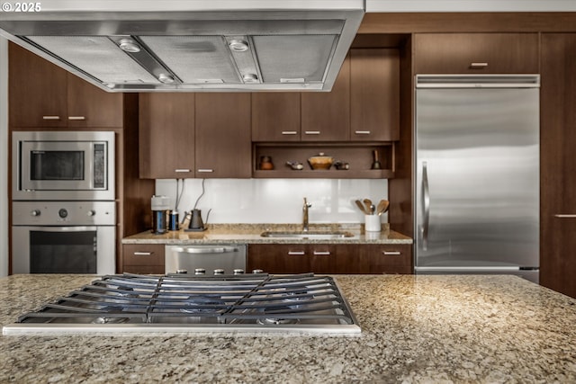 kitchen with light stone counters, a sink, built in appliances, and exhaust hood