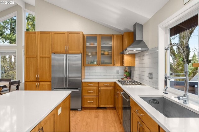 kitchen with vaulted ceiling, stainless steel appliances, wall chimney range hood, and brown cabinets