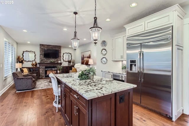 kitchen featuring a fireplace, hanging light fixtures, stainless steel built in fridge, dark brown cabinetry, and light wood-type flooring