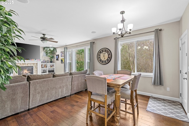 dining space with dark wood finished floors, a glass covered fireplace, ceiling fan with notable chandelier, and baseboards