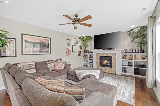 living room with dark wood-style floors, visible vents, a tiled fireplace, and a ceiling fan