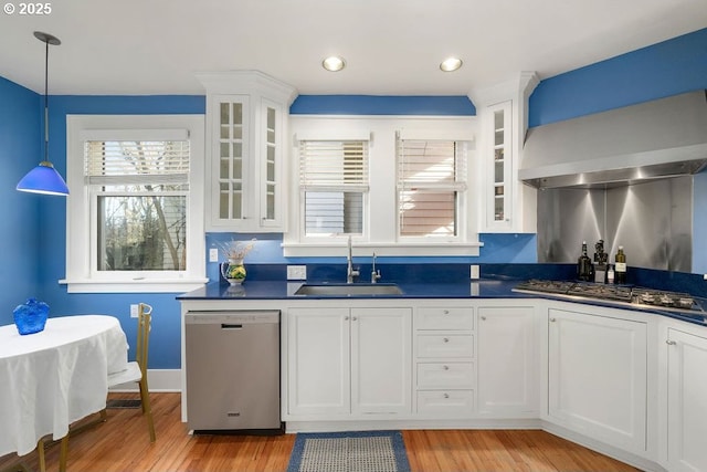 kitchen with dark countertops, appliances with stainless steel finishes, white cabinetry, a sink, and wall chimney range hood