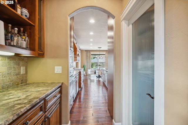 kitchen with arched walkways, brown cabinets, dark wood-type flooring, backsplash, and light stone counters