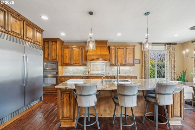 kitchen featuring a kitchen island with sink, a sink, custom range hood, brown cabinets, and built in appliances