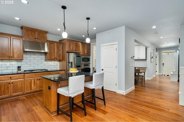 kitchen featuring a breakfast bar, under cabinet range hood, dark countertops, stainless steel appliances, and light wood finished floors