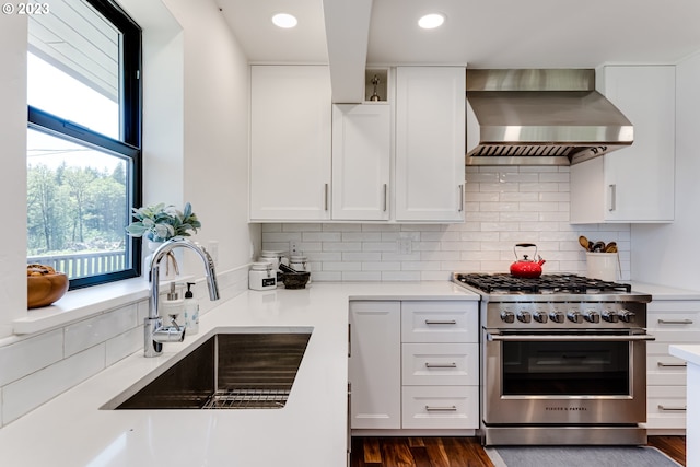 kitchen with light countertops, high end range, white cabinetry, wall chimney exhaust hood, and a sink