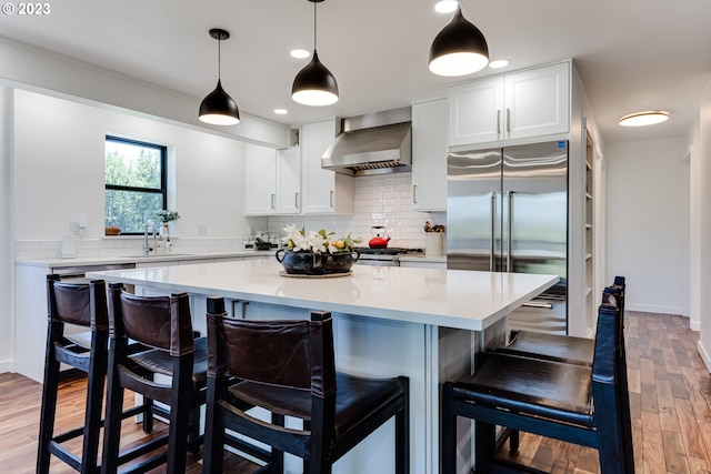 kitchen featuring a sink, light wood-style floors, wall chimney range hood, tasteful backsplash, and stainless steel built in refrigerator