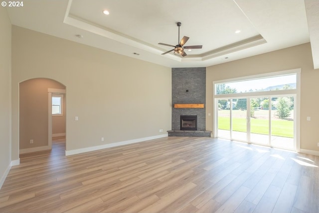 unfurnished living room with a raised ceiling, a stone fireplace, ceiling fan, and light hardwood / wood-style flooring