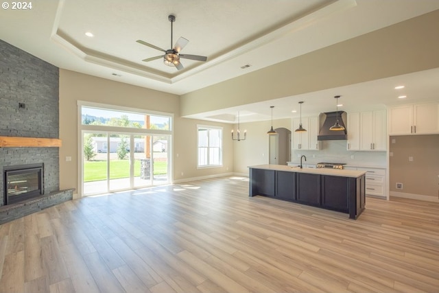 kitchen featuring custom range hood, a tray ceiling, a center island with sink, white cabinets, and ceiling fan with notable chandelier