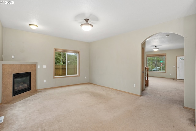 unfurnished living room featuring arched walkways, a tile fireplace, a wealth of natural light, and light colored carpet