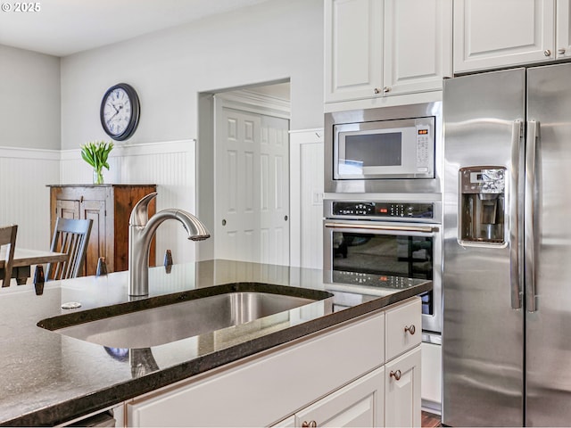 kitchen with stainless steel appliances, a sink, dark stone countertops, white cabinetry, and wainscoting