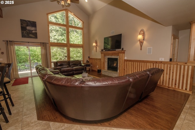 living area with a ceiling fan, wainscoting, light tile patterned floors, high vaulted ceiling, and a glass covered fireplace