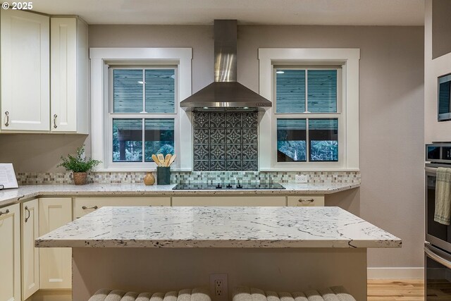 kitchen with black electric stovetop, light stone counters, oven, decorative backsplash, and wall chimney exhaust hood