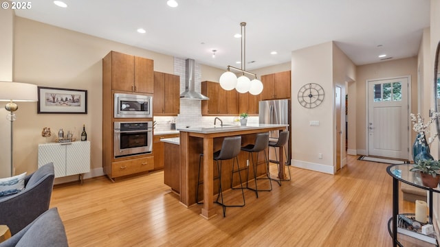 kitchen featuring brown cabinetry, appliances with stainless steel finishes, light countertops, a breakfast bar, and wall chimney exhaust hood