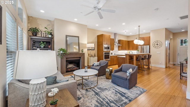 living room with a ceiling fan, visible vents, baseboards, light wood finished floors, and a glass covered fireplace