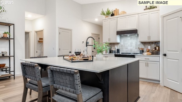 kitchen with light wood-style flooring, stainless steel range with electric cooktop, under cabinet range hood, a kitchen bar, and backsplash