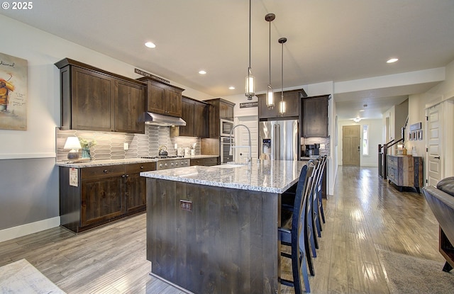 kitchen with backsplash, light stone countertops, under cabinet range hood, dark brown cabinetry, and appliances with stainless steel finishes