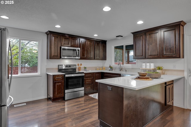 kitchen with dark brown cabinetry, dark wood finished floors, a wealth of natural light, a peninsula, and stainless steel appliances