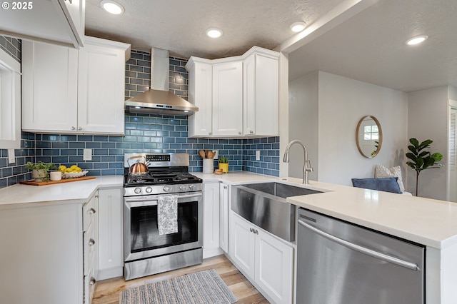 kitchen featuring stainless steel appliances, wall chimney range hood, a sink, a peninsula, and light wood finished floors