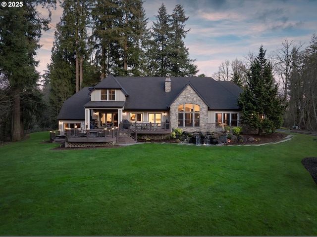 back of house at dusk featuring stone siding, a wooden deck, a chimney, and a yard