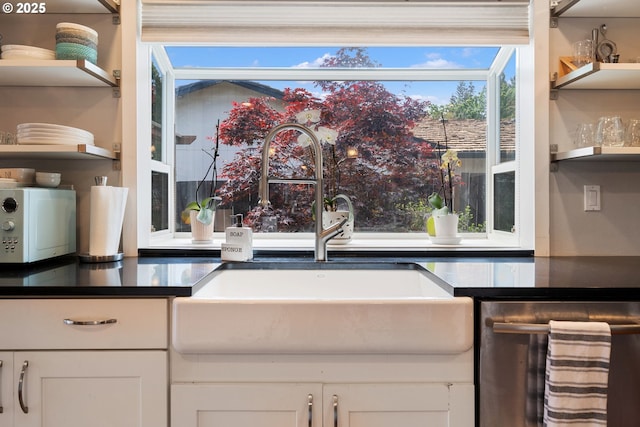 kitchen featuring stainless steel dishwasher, open shelves, dark countertops, white cabinetry, and a sink