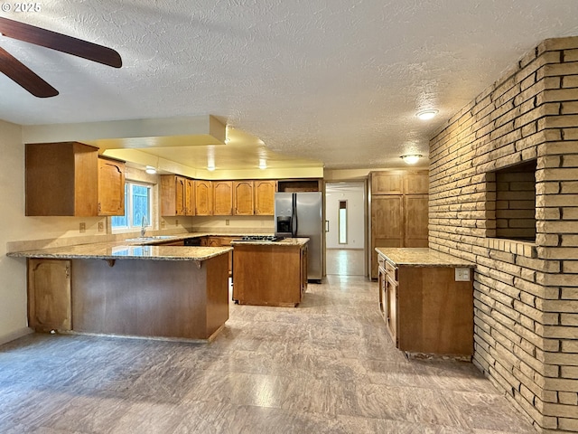 kitchen with a ceiling fan, a peninsula, brown cabinetry, stainless steel fridge with ice dispenser, and light stone countertops