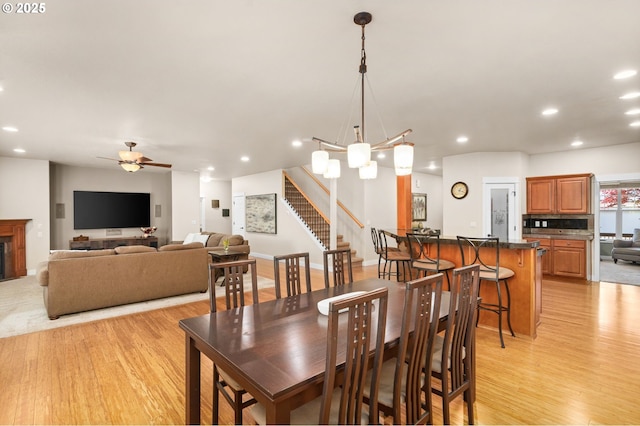 dining space featuring a fireplace, stairway, ceiling fan with notable chandelier, recessed lighting, and light wood-style flooring