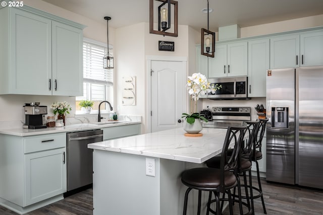 kitchen with light stone countertops, a sink, hanging light fixtures, appliances with stainless steel finishes, and a center island