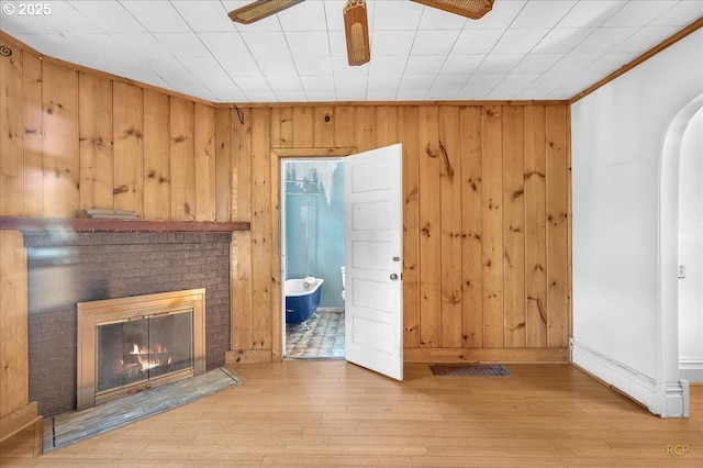 unfurnished living room with wooden walls, a ceiling fan, light wood-style floors, ornamental molding, and a brick fireplace