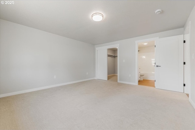 unfurnished bedroom with a closet, light carpet, baseboards, a walk in closet, and ensuite bath