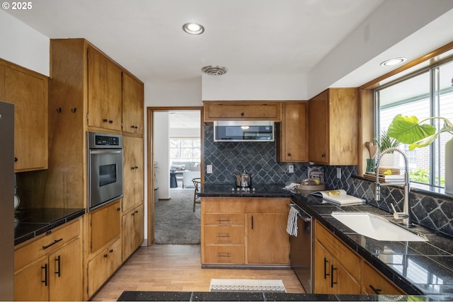 kitchen with stainless steel appliances, a sink, brown cabinetry, dark countertops, and tasteful backsplash