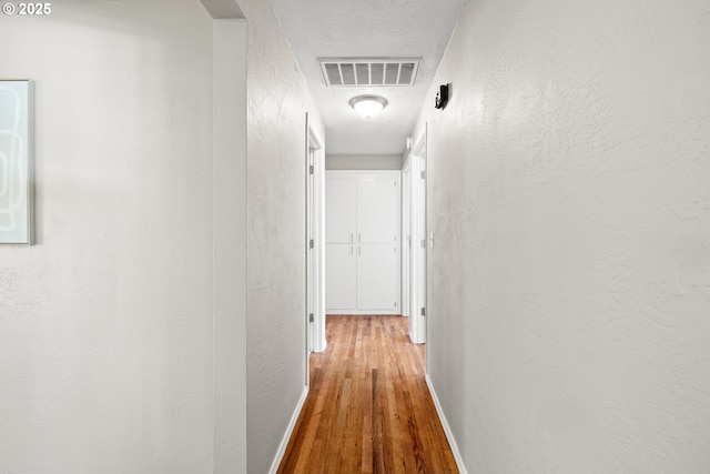 hall featuring wood finished floors, baseboards, visible vents, a textured ceiling, and a textured wall