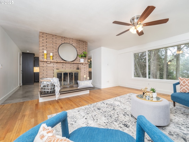 living room with ceiling fan, baseboards, a brick fireplace, and wood finished floors