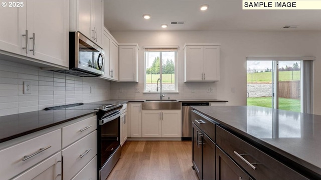 kitchen with dark countertops, appliances with stainless steel finishes, white cabinets, a sink, and light wood-type flooring